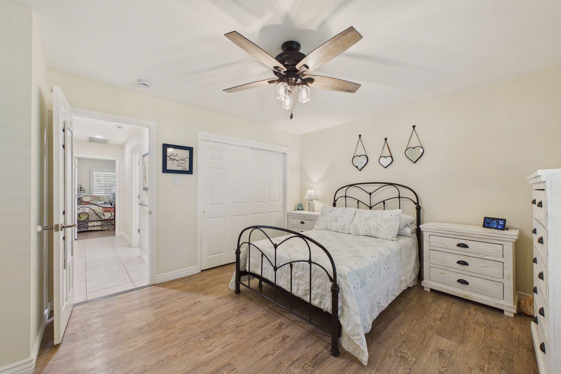 A bedroom with a black metal-framed bed, ceiling fan, white chest of drawers and wood-look flooring.