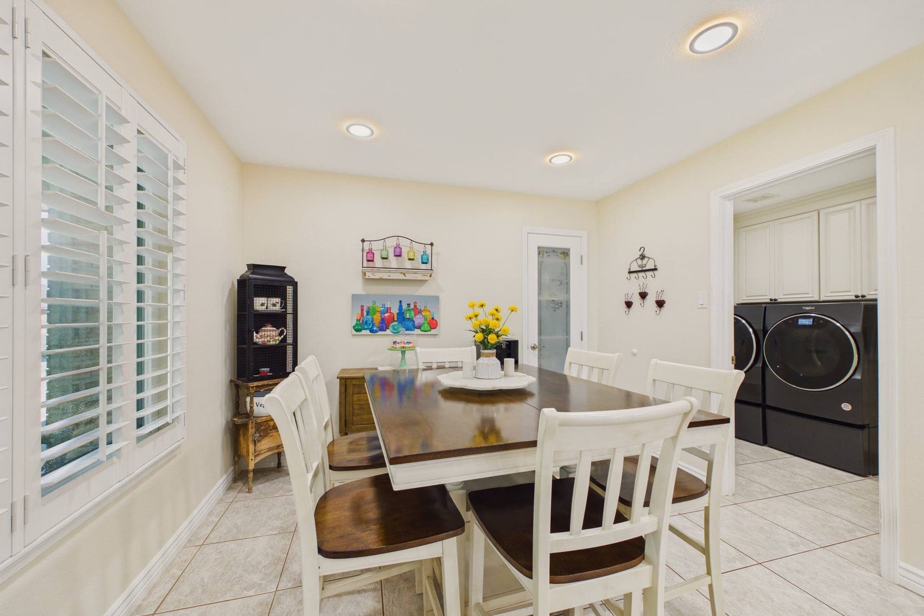 The dining area: a dark wood table with six cream chairs beside a shuttered window, opening to the laundry alcove.