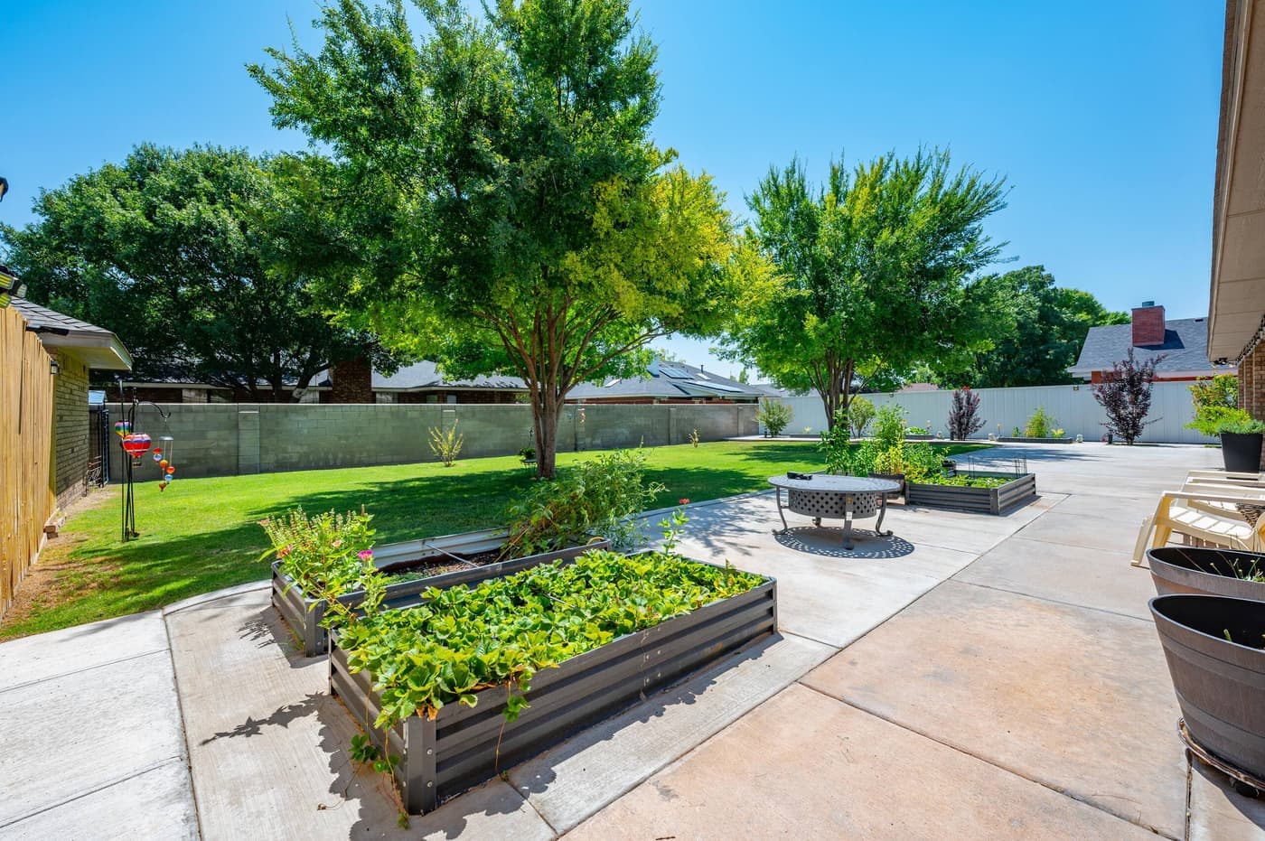 A round fire pit on the back patio beside raised vegetable beds, with mature trees and a block wall behind.