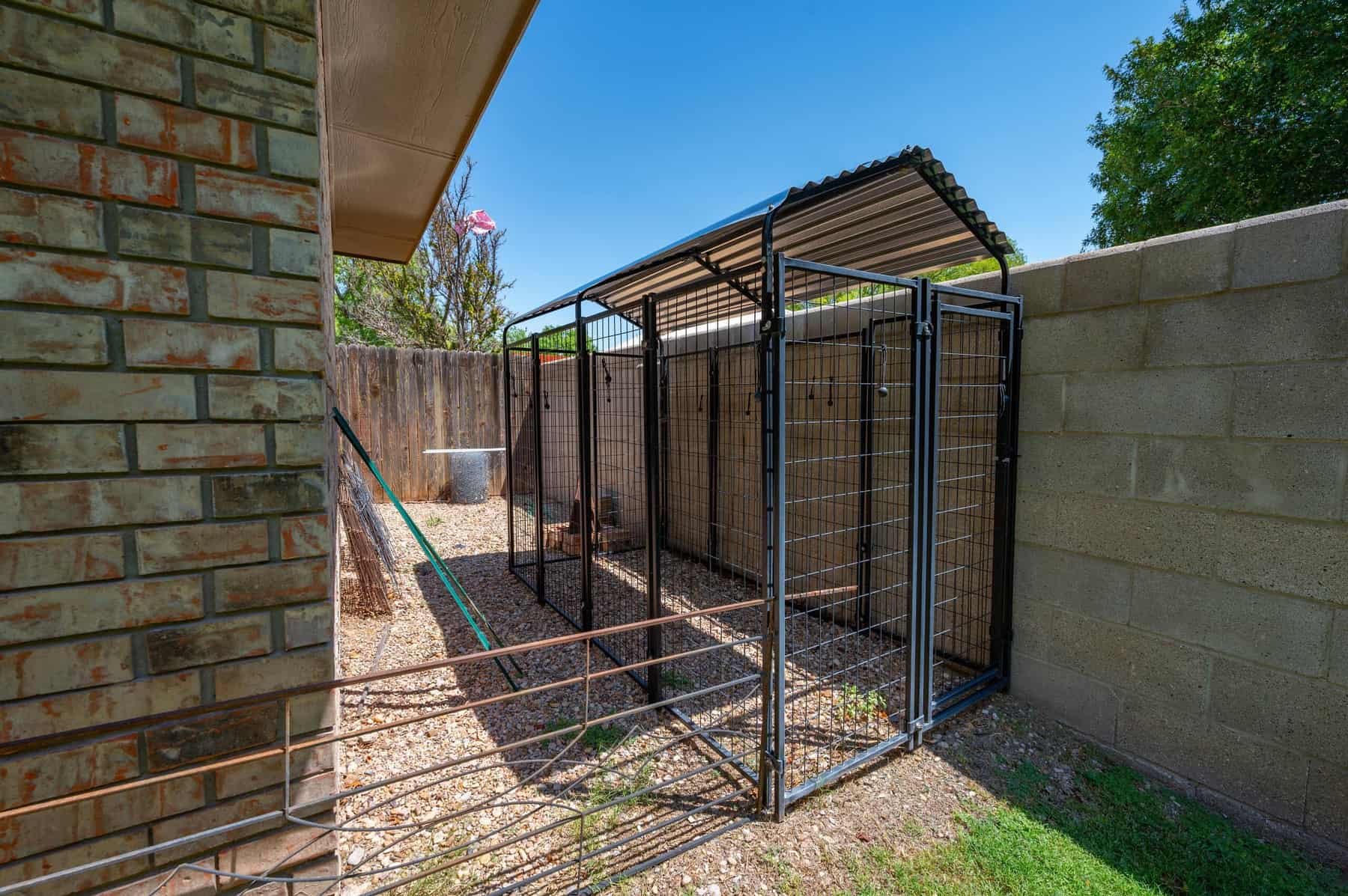 A covered wire kennel run on gravel along the side of the house, sheltered by the roof overhang.