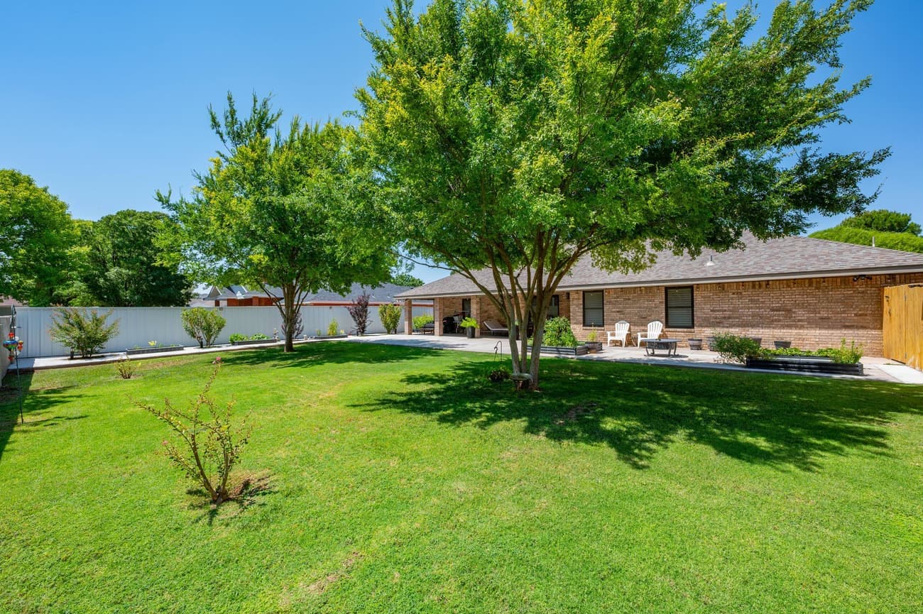 The back lawn, enclosed by a white privacy fence, with two mature shade trees and the covered patio along the back of the house.