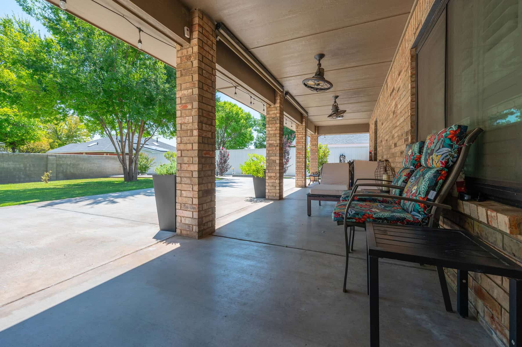 Patio seating in the shade of the covered porch, with brick columns and ceiling fans overhead and the lawn beyond.