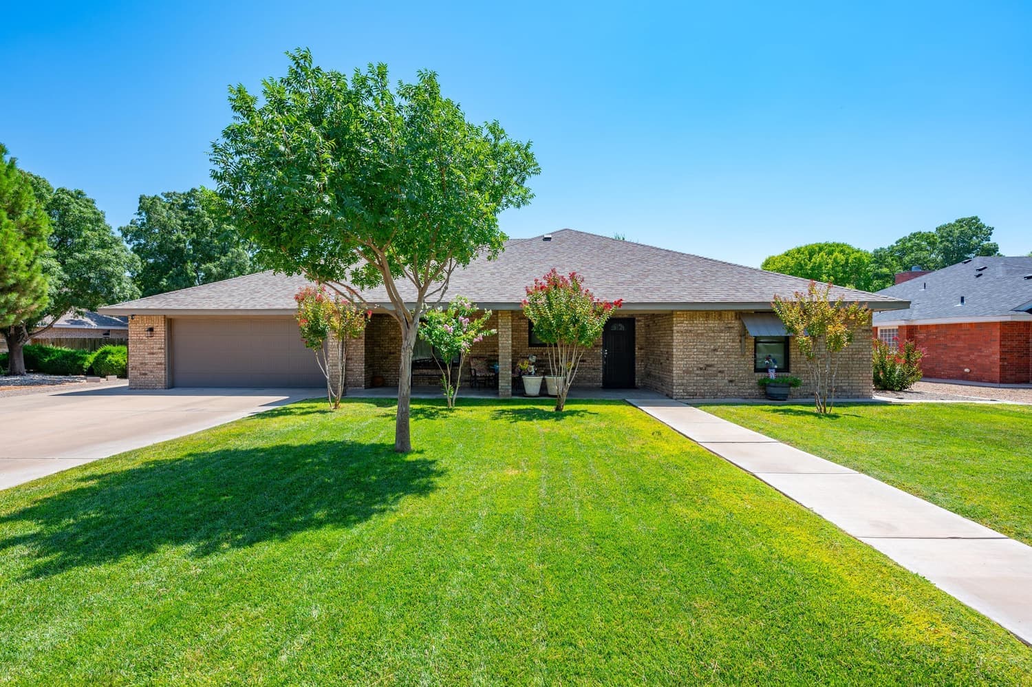 The front of Lana's Desert Haven: a single-story light-brick house with a mature shade tree, crepe myrtles in bloom and a watered green lawn under a clear New Mexico sky.