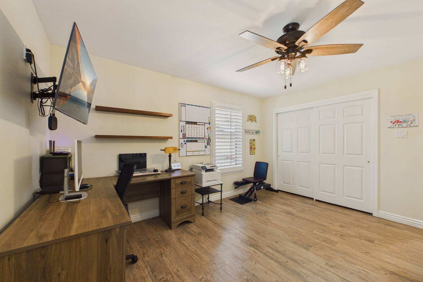 The dedicated workspace: an L-shaped desk with a monitor, a printer on a side table, wall shelving, a mounted television and a shuttered window.